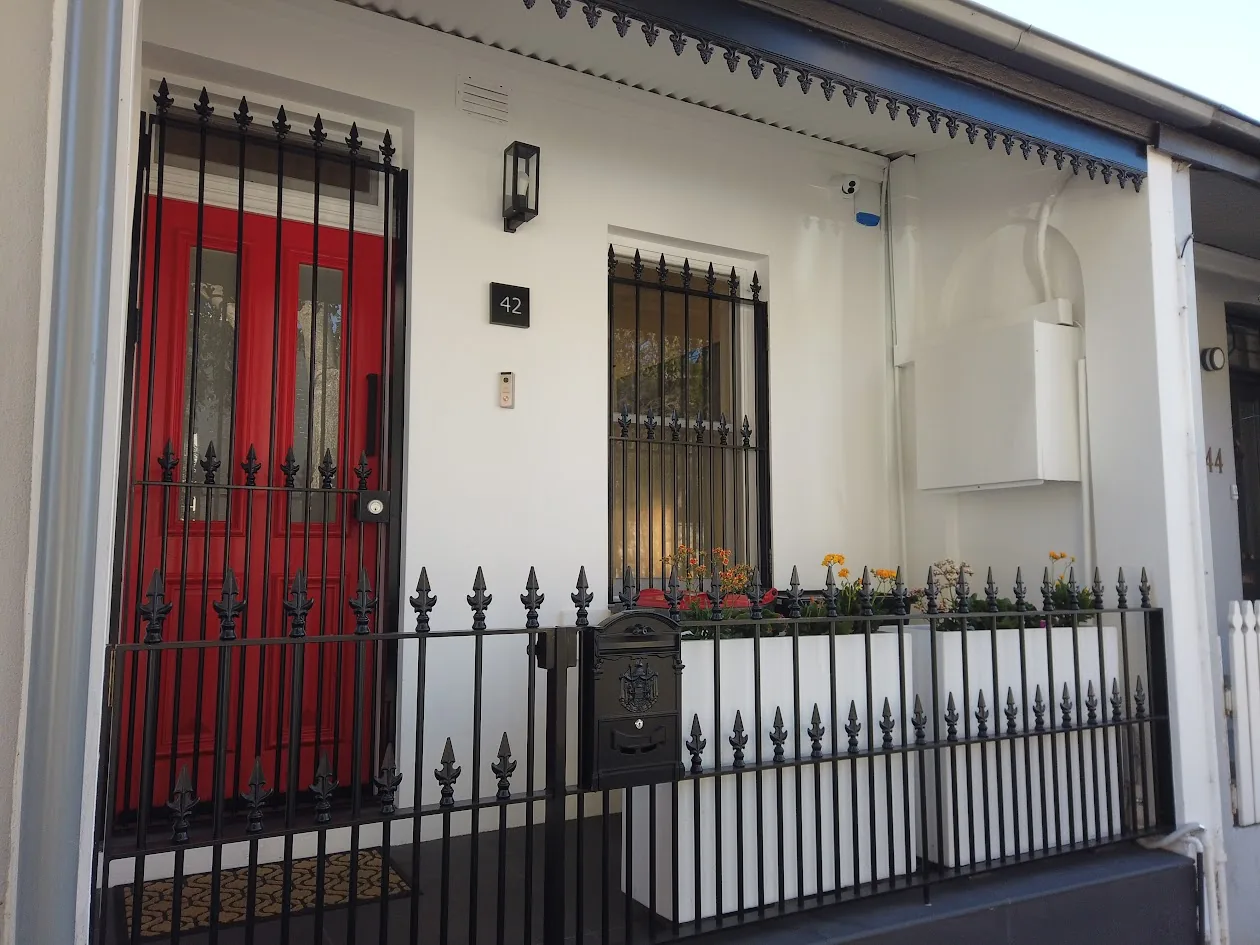Street frontage of a white-rendered Victorian terrace with red entry door, iron lacework fence and security bars on timber-framed windows