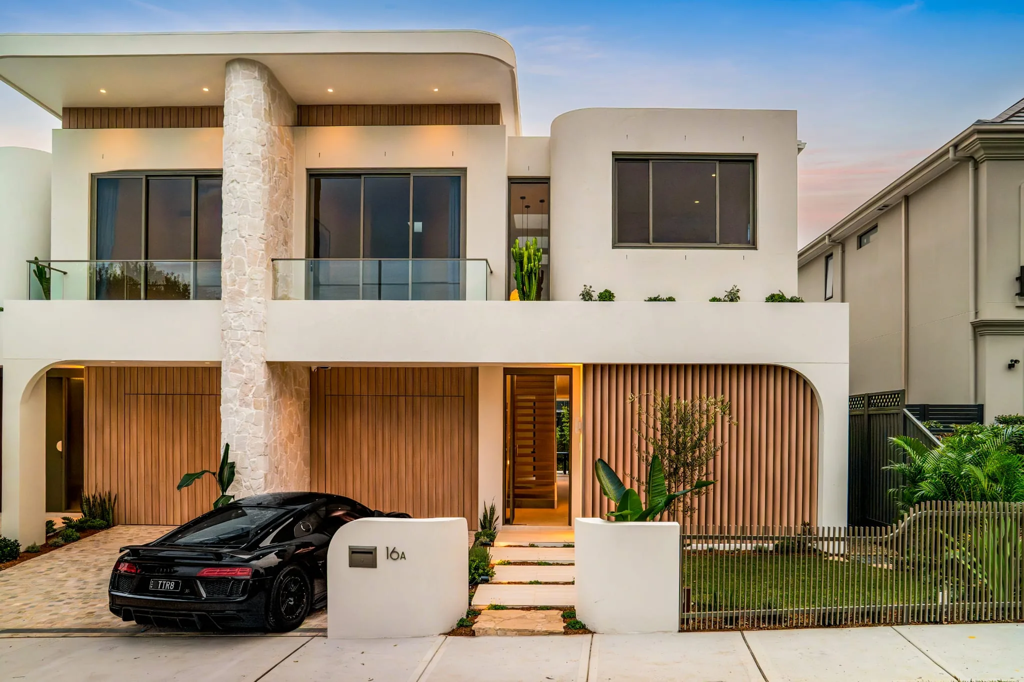Street-facing façade of the Concord Duplex at dusk — rendered white exterior, stone-clad central column, timber vertical battens, arched entry and upper-level glass balustrades