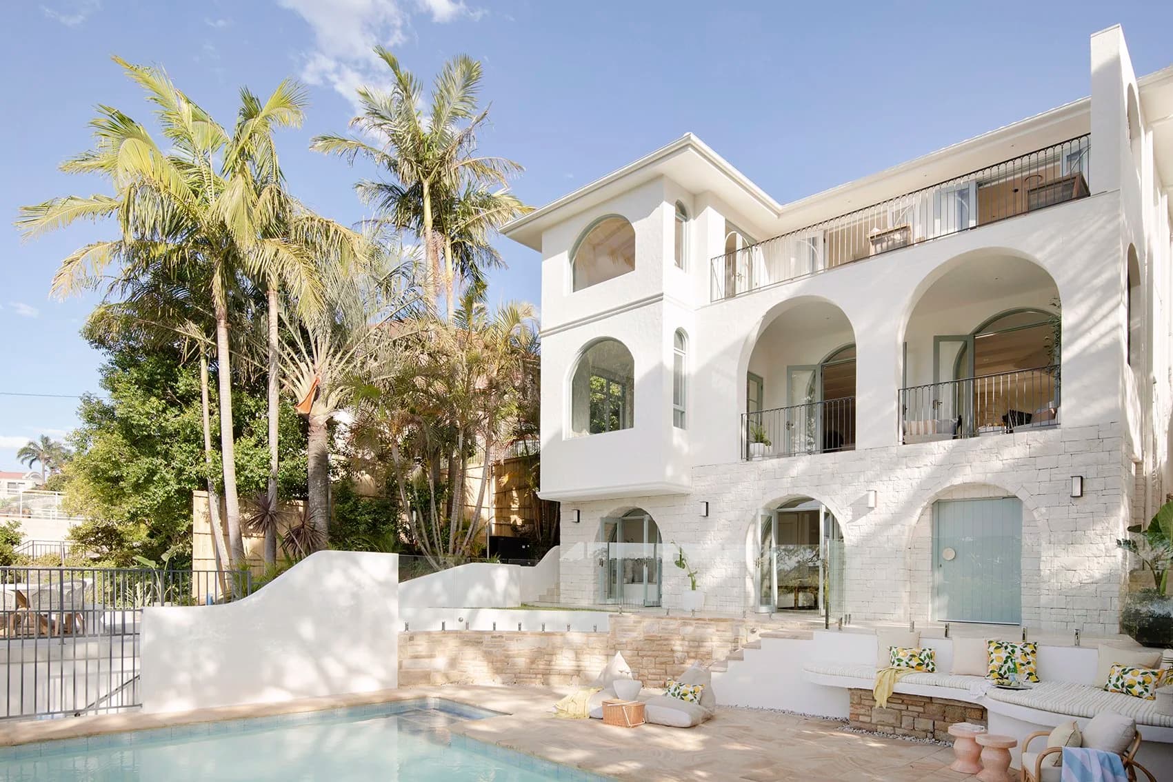 Rear exterior of a white rendered multi-storey Mosman home with arched windows, pool, palm trees and outdoor seating